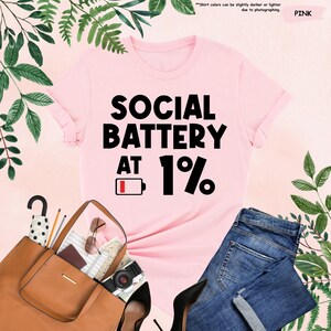 May include: A pink t-shirt with the text "SOCIAL BATTERY AT 1%" in black. The shirt is styled with a brown purse, sunglasses, a camera, and blue jeans. Green foliage surrounds the items.