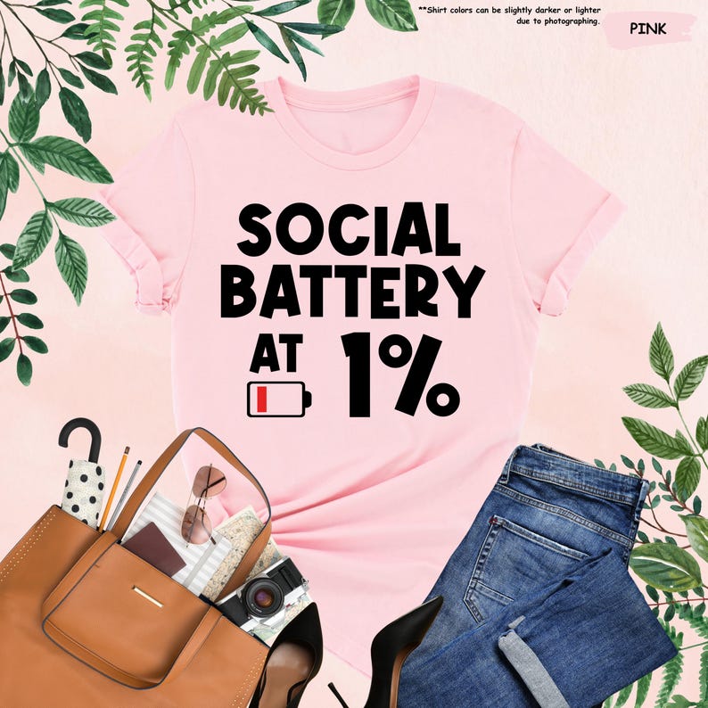 May include: A pink t-shirt with the text "SOCIAL BATTERY AT 1%" in black. The shirt is styled with a brown purse, sunglasses, a camera, and blue jeans. Green foliage surrounds the items.