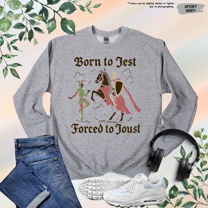 K&ouml;nnte beinhalten: Sportgraues Sweatshirt mit dem Text "Born to Jest Forced to Joust" und einer mittelalterlichen Grafik. Die Grafik zeigt einen Narren und einen Ritter zu Pferd. Das Sweatshirt wird mit Jeans, Turnschuhen und Kopfh&ouml;rern pr&auml;sentiert.