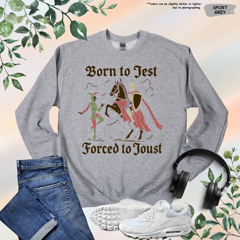 K&ouml;nnte beinhalten: Sportgraues Sweatshirt mit dem Text "Born to Jest Forced to Joust" und einer mittelalterlichen Grafik. Die Grafik zeigt einen Narren und einen Ritter zu Pferd. Das Sweatshirt wird mit Jeans, Turnschuhen und Kopfh&ouml;rern pr&auml;sentiert.
