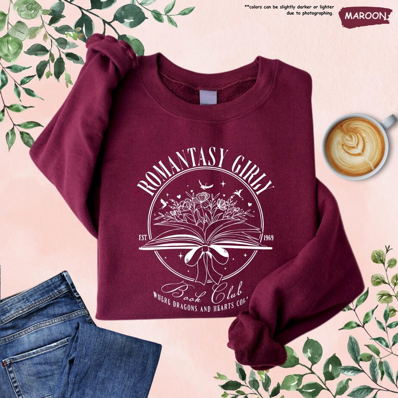 May include: A maroon sweatshirt with a white graphic design that says "Romantasy Girly Book Club Where Dragons and Hearts Collide" with a book, flowers, and birds. The text "Est 1969" is also included in the design.