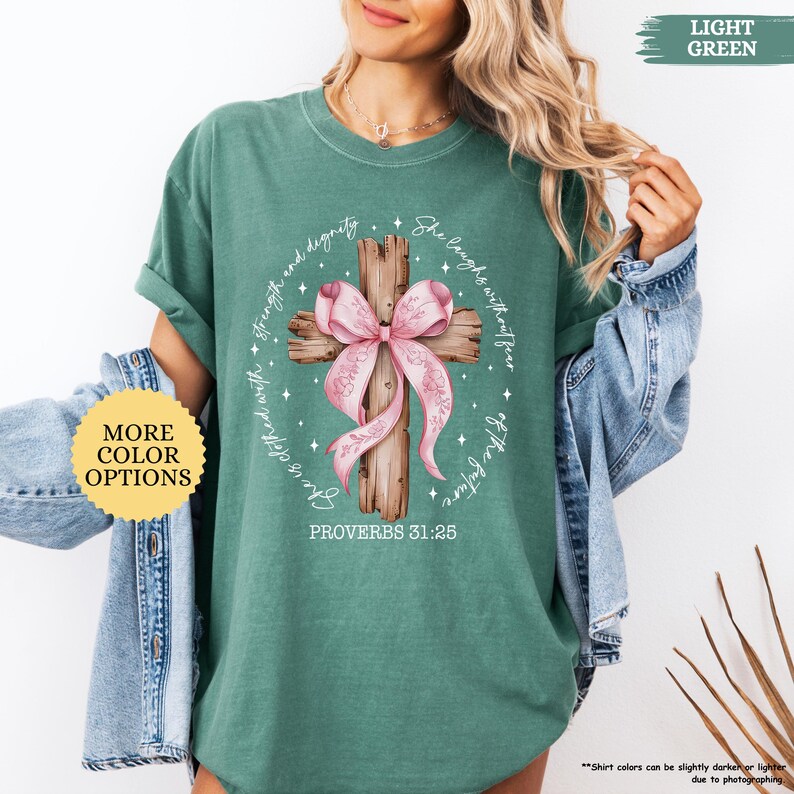 Op de afbeelding: Een lichtgroen T-shirt met een houten kruis en een roze strik. Het kruis is omringd door de woorden "Zij lacht zonder angst voor de toekomst" en "gekleed met kracht en waardigheid". De tekst "Spreuken 31:25" staat onder het kruis.