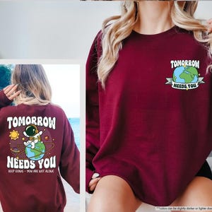 May include: A maroon sweatshirt featuring the text "TOMORROW NEEDS YOU" and an astronaut graphic. The back of the sweatshirt has a larger graphic with the same text and an astronaut on a planet. The phrase "KEEP GOING - YOU ARE NOT ALONE" is also included.