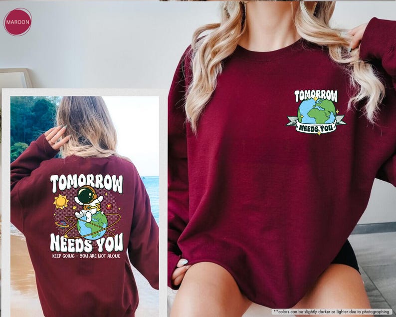 May include: A maroon sweatshirt featuring the text "TOMORROW NEEDS YOU" and an astronaut graphic. The back of the sweatshirt has a larger graphic with the same text and an astronaut on a planet. The phrase "KEEP GOING - YOU ARE NOT ALONE" is also included.