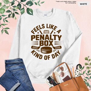May include: White sweatshirt with the text "FEELS LIKE A PENALTY BOX KIND OF DAY" in brown. The design includes hockey sticks, a puck, and a net. The sweatshirt is displayed with blue jeans and a brown purse.