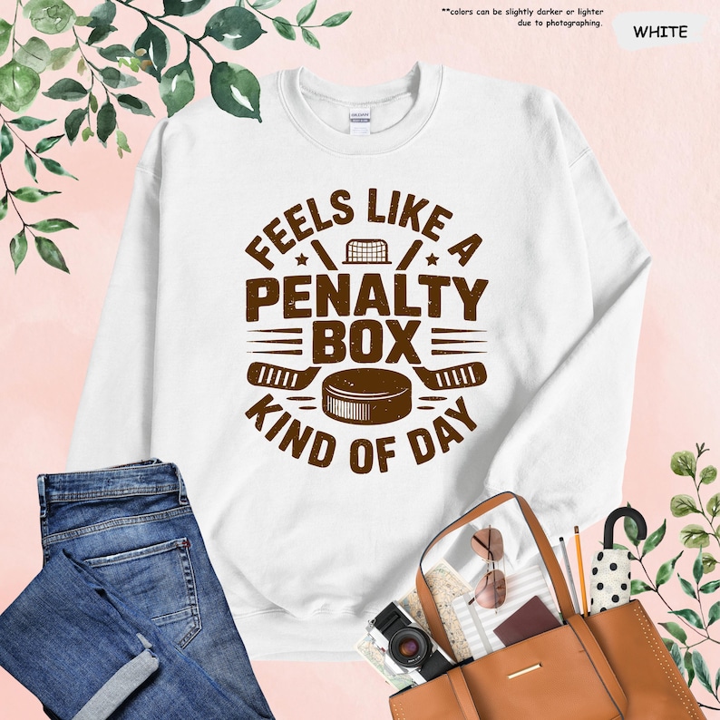 May include: White sweatshirt with the text "FEELS LIKE A PENALTY BOX KIND OF DAY" in brown. The design includes hockey sticks, a puck, and a net. The sweatshirt is displayed with blue jeans and a brown purse.