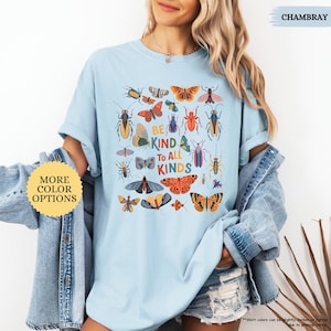 May include: Pale blue t-shirt with a vibrant insect and butterfly design. The shirt displays the words "BE KIND TO ALL KINDS". The garment is part of a collection with more colour options. The fabric is chambray.