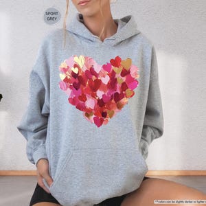Pu&ograve; includere: Felpa con cappuccio grigio sportivo con un grande design a cuore realizzato con pennellate rosse, rosa e oro. La felpa con cappuccio ha un cappuccio con coulisse e una tasca frontale. Il testo "SPORT GREY" &egrave; in un cerchio in alto.
