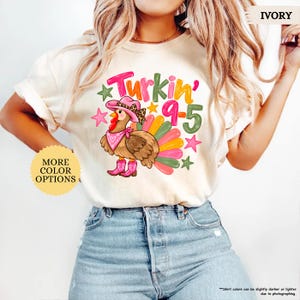 May include: An ivory t-shirt featuring a graphic of a turkey wearing a leopard print cowboy hat, a pink bandana, and pink cowboy boots. The text reads "Turkin' 9-5" in colourful letters, surrounded by pink and green stars and a colourful tail.