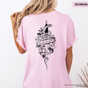 May include: Pale pink t-shirt with a black graphic on the back. The graphic includes a sword, snake, flowers, and the text "you do not yield" on a banner. The word "BLOSSOM" is printed at the top.