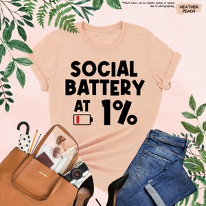 May include: A peach-colored t-shirt with the text "SOCIAL BATTERY AT 1%" in black. The shirt is displayed with a brown purse, blue jeans, and black heels. The words "HEATHER PEACH" are in the upper right corner.