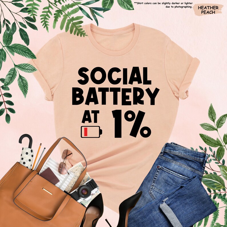 May include: A peach-colored t-shirt with the text "SOCIAL BATTERY AT 1%" in black. The shirt is displayed with a brown purse, blue jeans, and black heels. The words "HEATHER PEACH" are in the upper right corner.