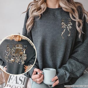 May include: Dark heather gray sweatshirt with a decorative bow and deer antlers design. The bow has a brown and tan animal print. The back of the sweatshirt has a larger design with a deer, hunting blind, and more antlers.