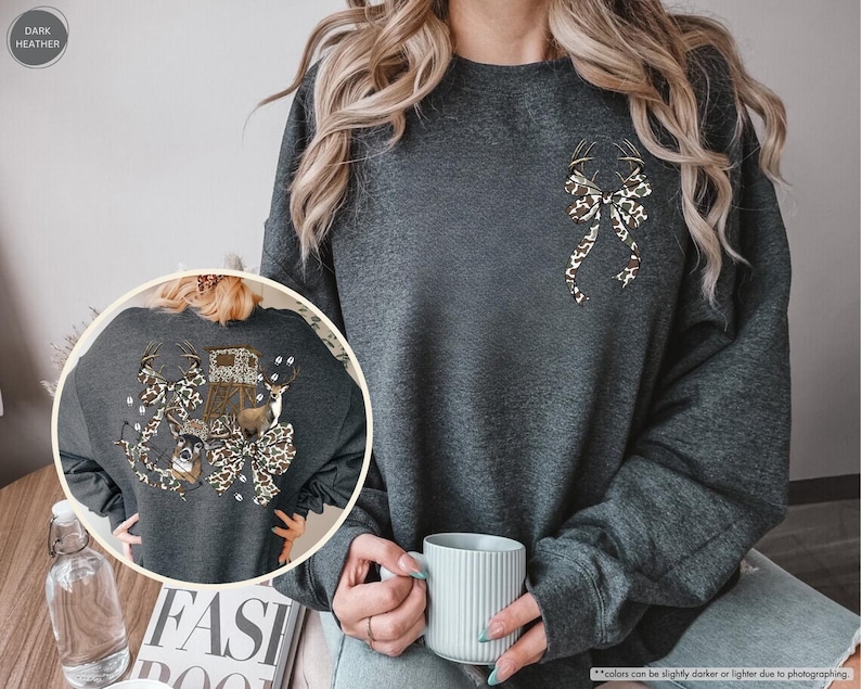 May include: Dark heather gray sweatshirt with a decorative bow and deer antlers design. The bow has a brown and tan animal print. The back of the sweatshirt has a larger design with a deer, hunting blind, and more antlers.