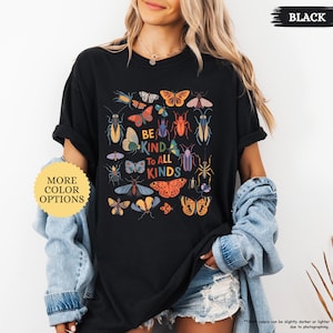 May include: Black t-shirt featuring a vibrant insect and butterfly design. The words "BE KIND TO ALL KINDS" are printed in the center. A yellow and white circular button with the text "MORE COLOR OPTIONS" is on the left.
