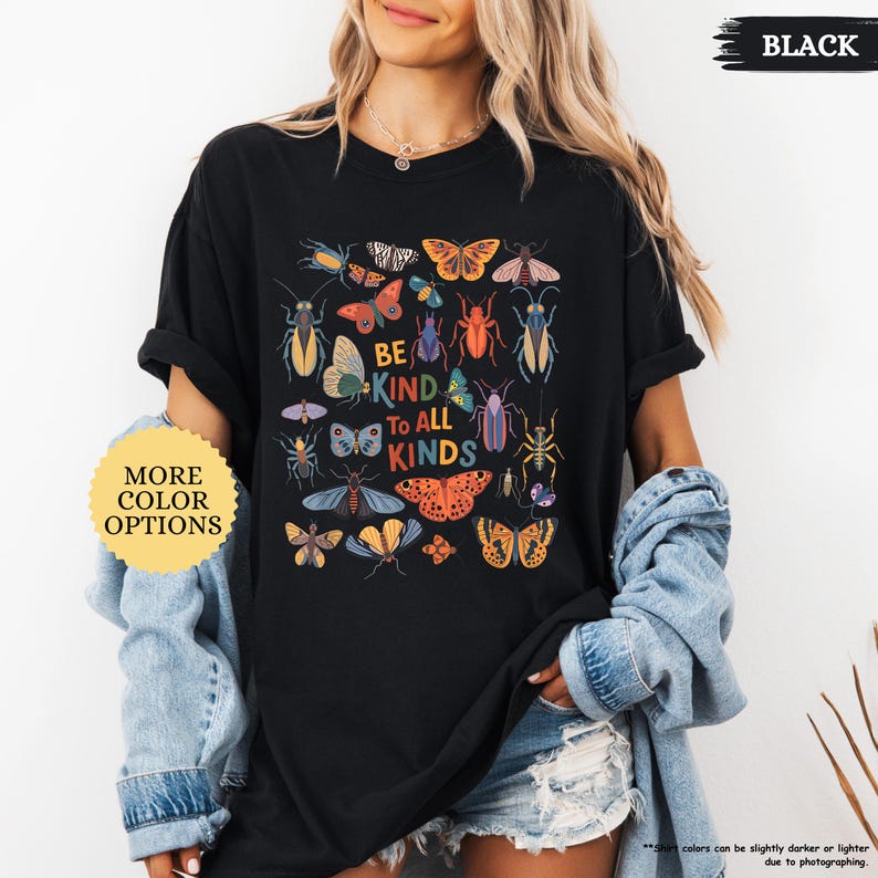 May include: Black t-shirt featuring a vibrant insect and butterfly design. The words "BE KIND TO ALL KINDS" are printed in the center. A yellow and white circular button with the text "MORE COLOR OPTIONS" is on the left.