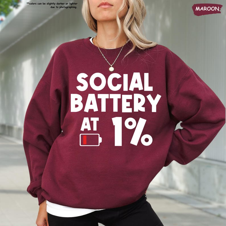 May include: Maroon sweatshirt with the text "SOCIAL BATTERY AT 1%" in white. The design includes a low battery icon. The sweatshirt is a solid maroon color.
