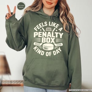 May include: Olive green hoodie with a front graphic that reads "FEELS LIKE A PENALTY BOX KIND OF DAY" in white. The design includes hockey-related imagery such as a net, hockey sticks, and a puck. The hoodie has a drawstring hood and a front pocket.