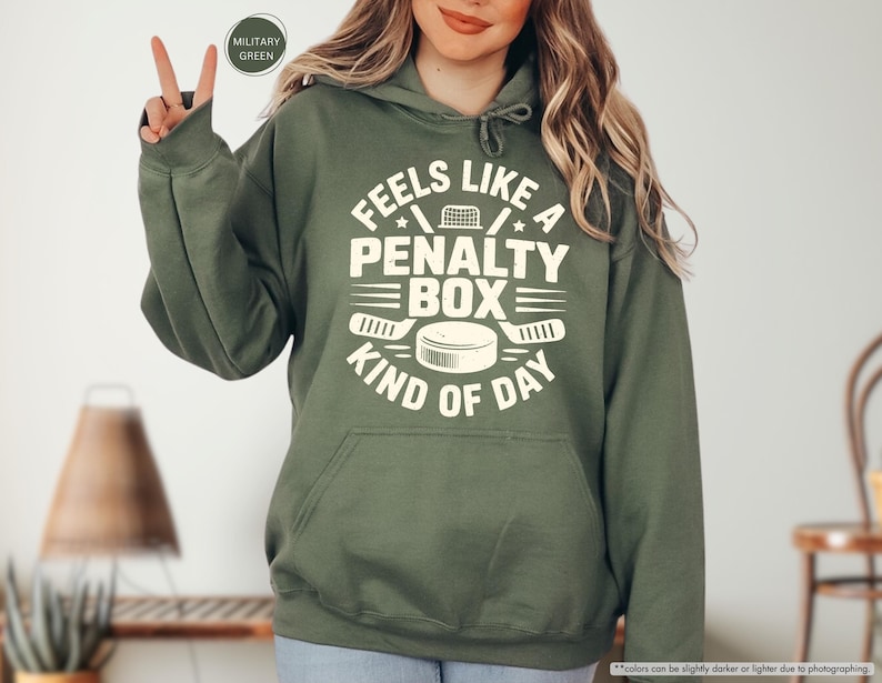 May include: Olive green hoodie with a front graphic that reads "FEELS LIKE A PENALTY BOX KIND OF DAY" in white. The design includes hockey-related imagery such as a net, hockey sticks, and a puck. The hoodie has a drawstring hood and a front pocket.