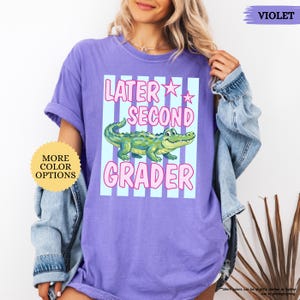 May include: A violet t-shirt with the text "LATER SECOND GRADER" in pink and white, featuring a green alligator illustration. The shirt has a light blue striped background and a "MORE COLOR OPTIONS" button.