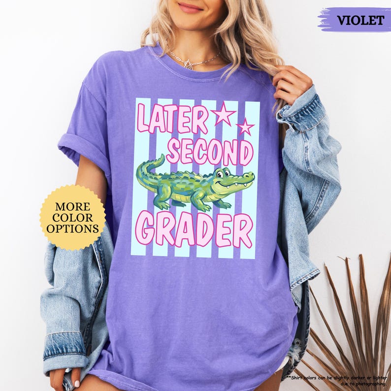 May include: A violet t-shirt with the text "LATER SECOND GRADER" in pink and white, featuring a green alligator illustration. The shirt has a light blue striped background and a "MORE COLOR OPTIONS" button.