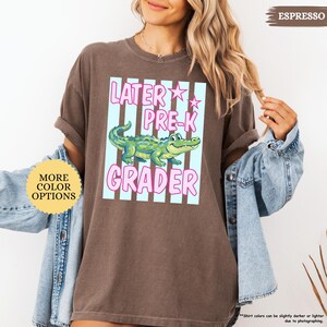May include: Espresso-colored t-shirt with a graphic that reads "Later Pre-K Grader." The design features a cartoon alligator in a light blue and pink striped box. The shirt also has a "More Color Options" button.