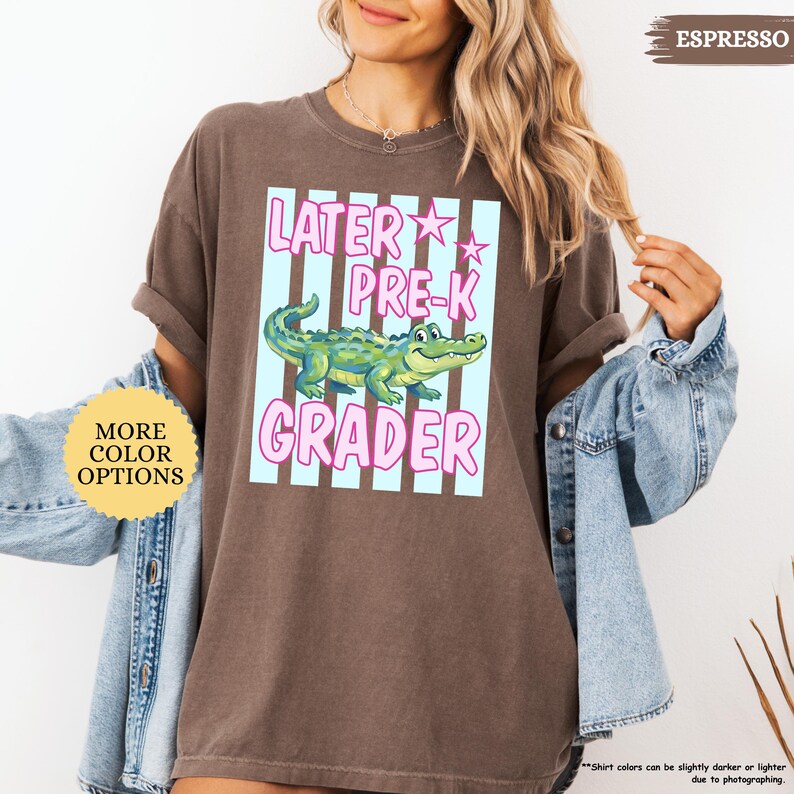 May include: Espresso-colored t-shirt with a graphic that reads "Later Pre-K Grader." The design features a cartoon alligator in a light blue and pink striped box. The shirt also has a "More Color Options" button.