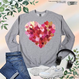 Pu&ograve; includere: Felpa grigio sport con un design a cuore composto da cuori rossi, rosa e oro. La felpa &egrave; mostrata con jeans blu, scarpe da ginnastica bianche e cuffie nere. Il testo "Sport Grey" &egrave; visibile.
