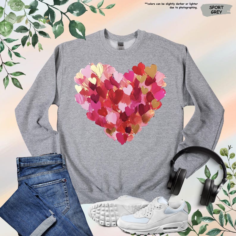 Pu&ograve; includere: Felpa grigio sport con un design a cuore composto da cuori rossi, rosa e oro. La felpa &egrave; mostrata con jeans blu, scarpe da ginnastica bianche e cuffie nere. Il testo "Sport Grey" &egrave; visibile.
