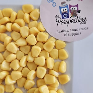 May include: A close-up shot of a white plate filled with realistic faux corn kernels. The kernels are a vibrant yellow color. A circular sticker with the text "Prop Chef" and "Two Perspectives Studio" is visible.