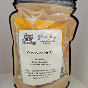 May include: A brown paper bag with a clear plastic window, shaped like a mason jar, containing a peach cobbler kit. The kit includes 6 faux peaches and 2 - 1oz Model Magic packs. The bag has a label that says "Prop Chef Realistic Faux Foods & Supplies" and "Peach Cobbler Kit". The label also includes instructions for the kit, which are available on Facebook at the Two Perspectives Studio page. The live tutorial was done on September 10, 2024.