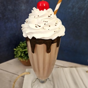 K&ouml;nnte beinhalten: Ein hohes Glas gef&uuml;llt mit einem Schokoladen-Milchshake, der mit Schlagsahne, Schokoladenstreuseln und einer roten Kirsche garniert ist. Ein brauner und wei&szlig;er Strohhalm befindet sich im Glas.