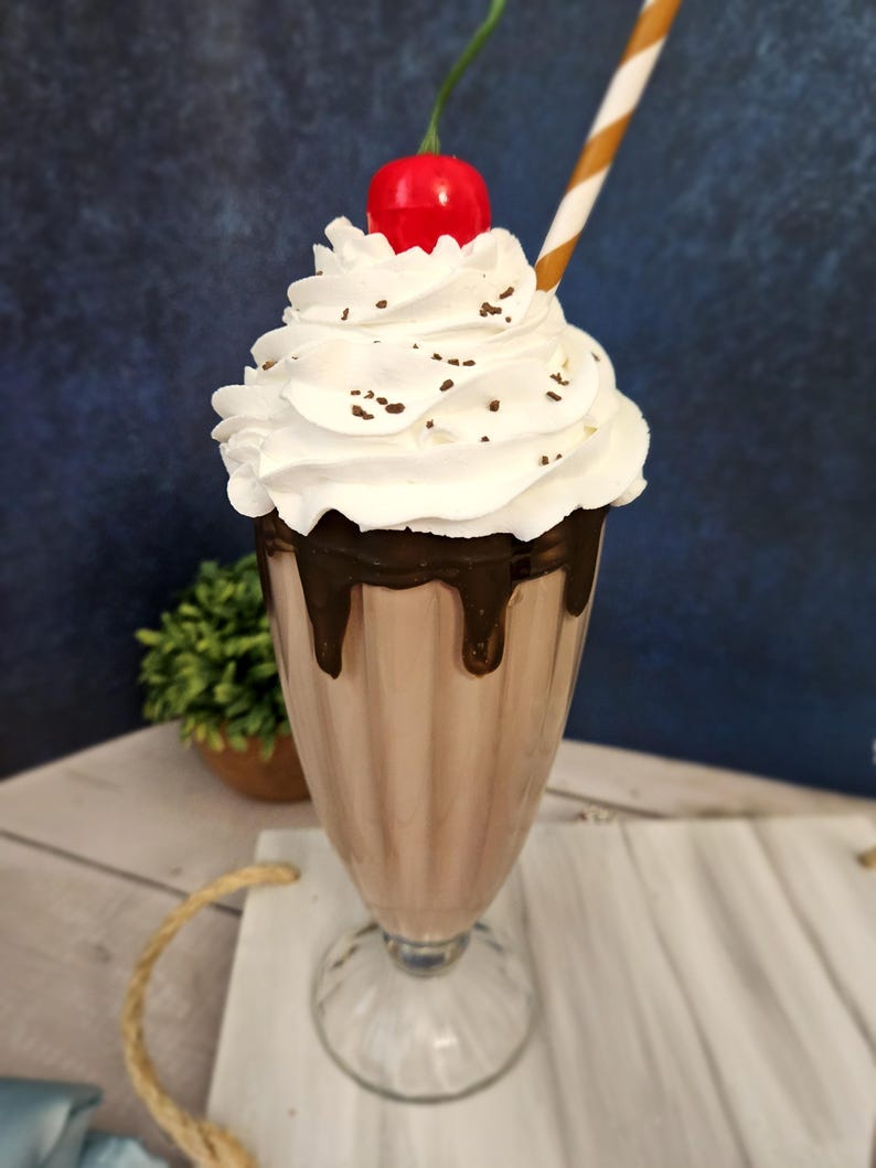 K&ouml;nnte beinhalten: Ein hohes Glas gef&uuml;llt mit einem Schokoladen-Milchshake, der mit Schlagsahne, Schokoladenstreuseln und einer roten Kirsche garniert ist. Ein brauner und wei&szlig;er Strohhalm befindet sich im Glas.
