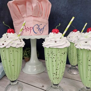 K&ouml;nnte beinhalten: Vier gr&uuml;ne Minz-Schokoladen-Milchshakes mit Schlagsahne und einer Kirsche obenauf. Jeder Milchshake hat einen gestreiften Strohhalm und befindet sich in einem klaren Glas mit breiter Basis. Ein rosa Kochhut mit dem Text "Prop Chef" steht auf einem wei&szlig;en Kuchenst&auml;nder hinter den Milchshakes.
