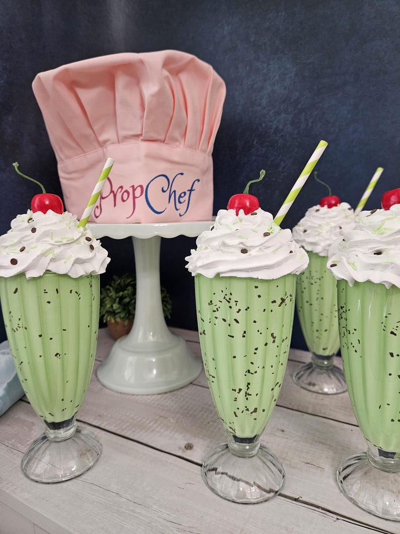 K&ouml;nnte beinhalten: Vier gr&uuml;ne Minz-Schokoladen-Milchshakes mit Schlagsahne und einer Kirsche obenauf. Jeder Milchshake hat einen gestreiften Strohhalm und befindet sich in einem klaren Glas mit breiter Basis. Ein rosa Kochhut mit dem Text "Prop Chef" steht auf einem wei&szlig;en Kuchenst&auml;nder hinter den Milchshakes.