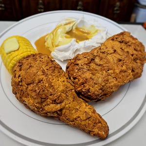 May include: A plate of food featuring fried chicken, corn on the cob with butter, and a dollop of whipped cream with a sauce. The fried chicken has a crispy, golden-brown exterior. The corn is yellow, and the whipped cream is white.