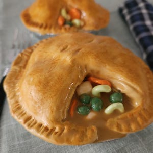 May include: Two golden-brown, savory pot pies with visible fillings of peas, carrots, and other vegetables. The pies sit on a light green surface, with a blue and black checkered cloth in the background. The pies have a flaky crust.