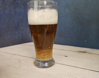 Puede incluir: Un vaso alto y transparente de cerveza con una espesa espuma blanca. La cerveza es de color ámbar oscuro, con una capa de sedimento dorado en el fondo. El vaso está sobre una superficie de madera clara.
