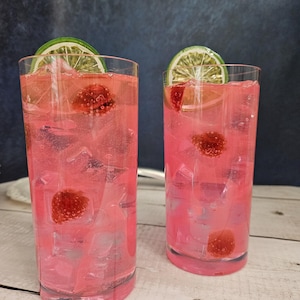 May include: Two tall glasses filled with pink lemonade, ice cubes, and raspberries. Each glass is topped with a lime wedge.