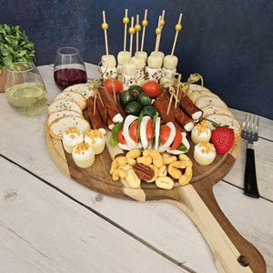 May include: A wooden serving board with a long handle, filled with a variety of snacks, including cheese cubes, crackers, nuts, and fruit. The board is decorated with small, white cupcakes and a few sprigs of greenery.