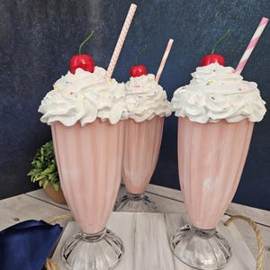 K&ouml;nnte beinhalten: Drei rosa Milchshakes mit Schlagsahne, Streuseln und Kirschen obenauf. Jeder Milchshake hat einen rosa-wei&szlig; gestreiften Strohhalm. Die Milchshakes stehen auf einem wei&szlig;en Holztablett mit einem Seilgriff.