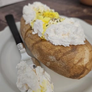 May include: A baked potato topped with whipped cream and a yellow sauce, garnished with herbs, sits on a white plate. A fork with a black handle rests beside the potato. A small dollop of the topping is on the plate.