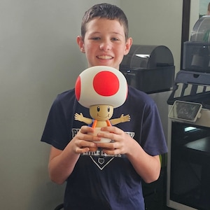May include: A young person wearing a blue shirt with a baseball logo and black shorts is holding a white and red mushroom character with a red dot on top. The character is from the Super Mario video game series.