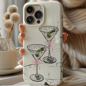May include: White iPhone case featuring a design of two martini glasses with olives and pink ribbons. The case has a camera lens cutout and a white background.