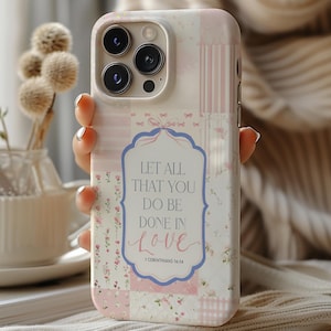 May include: A phone case with a patchwork design in pink, white, and cream colors. The case features a floral pattern and the text "LET ALL THAT YOU DO BE DONE IN LOVE" in a decorative frame. The case has a camera cutout and buttons.