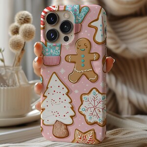 May include: Pink phone case with a Christmas cookie design. The case features gingerbread men, snowflakes, and Christmas trees.