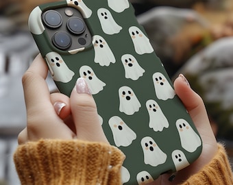 May include: A dark green phone case featuring a pattern of white cartoon ghosts with black eyes and mouths. The case is held in front of a blurred outdoor background, suggesting a fall or Halloween theme.