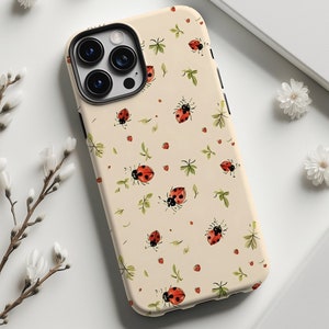 May include: A beige phone case featuring a repeating pattern of red ladybugs and green leaves. The case has a black camera module and a black border. White flowers and a white notebook are in the background.