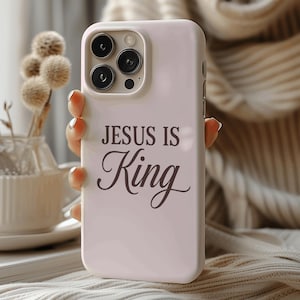 Può includere: Una custodia per smartphone rosa chiaro con un'apertura per la fotocamera a tripla lente e il testo marrone scuro "JESUS IS King" stampato sul retro. La custodia ha una finitura liscia e copre il retro e i lati del dispositivo mobile.