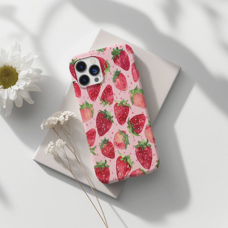 May include: A pink phone case featuring a pattern of red strawberries. The case has a camera lens cutout and a white background.